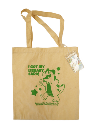 A cotton totebag with a luggage tag attached to it. The totebag has a drawing of a dinosaur on it, and reads "I got my library card!"