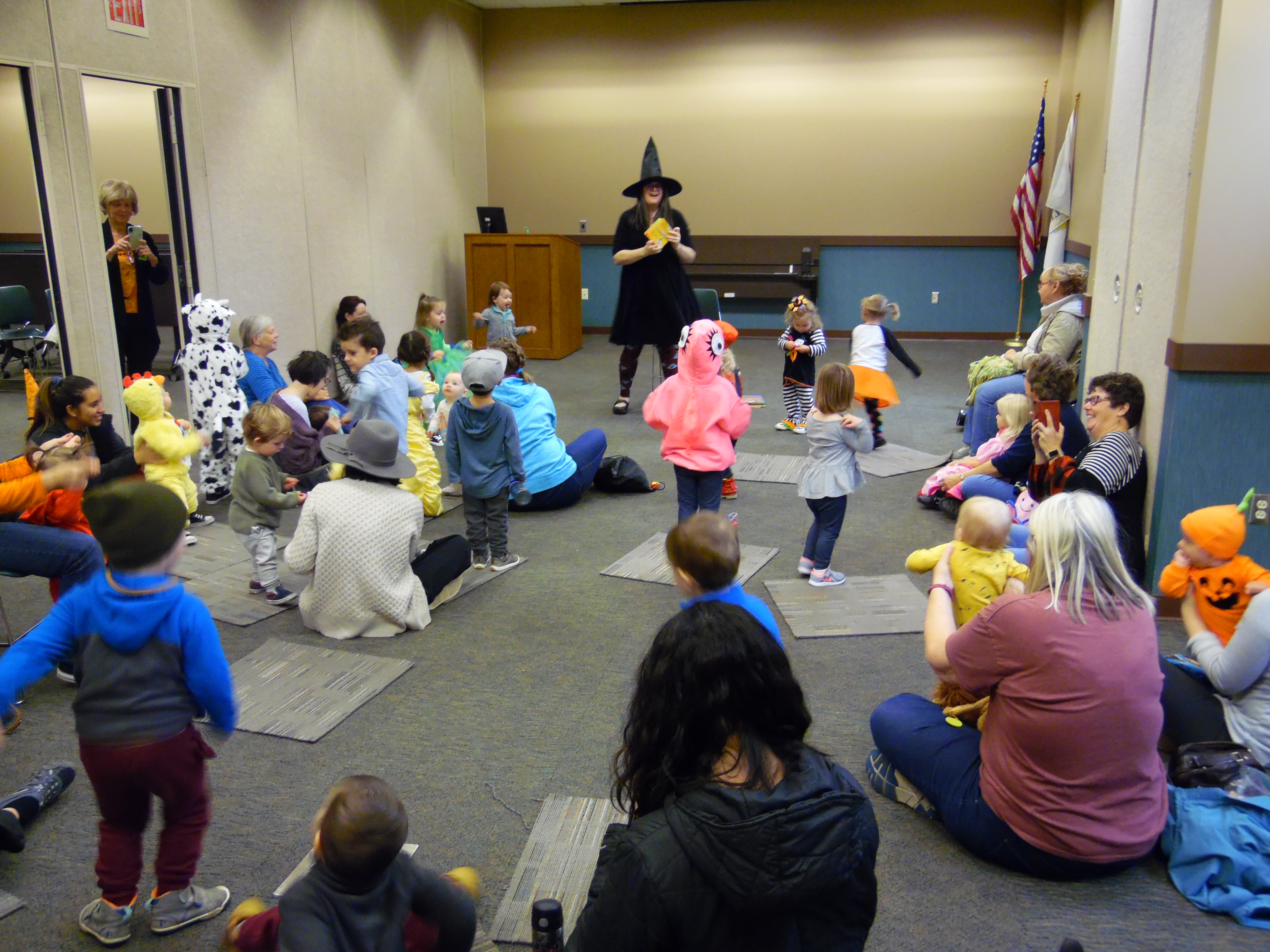 A woman dressed in a black dress and black witch's hat reads to a group of parents and babies in costume.