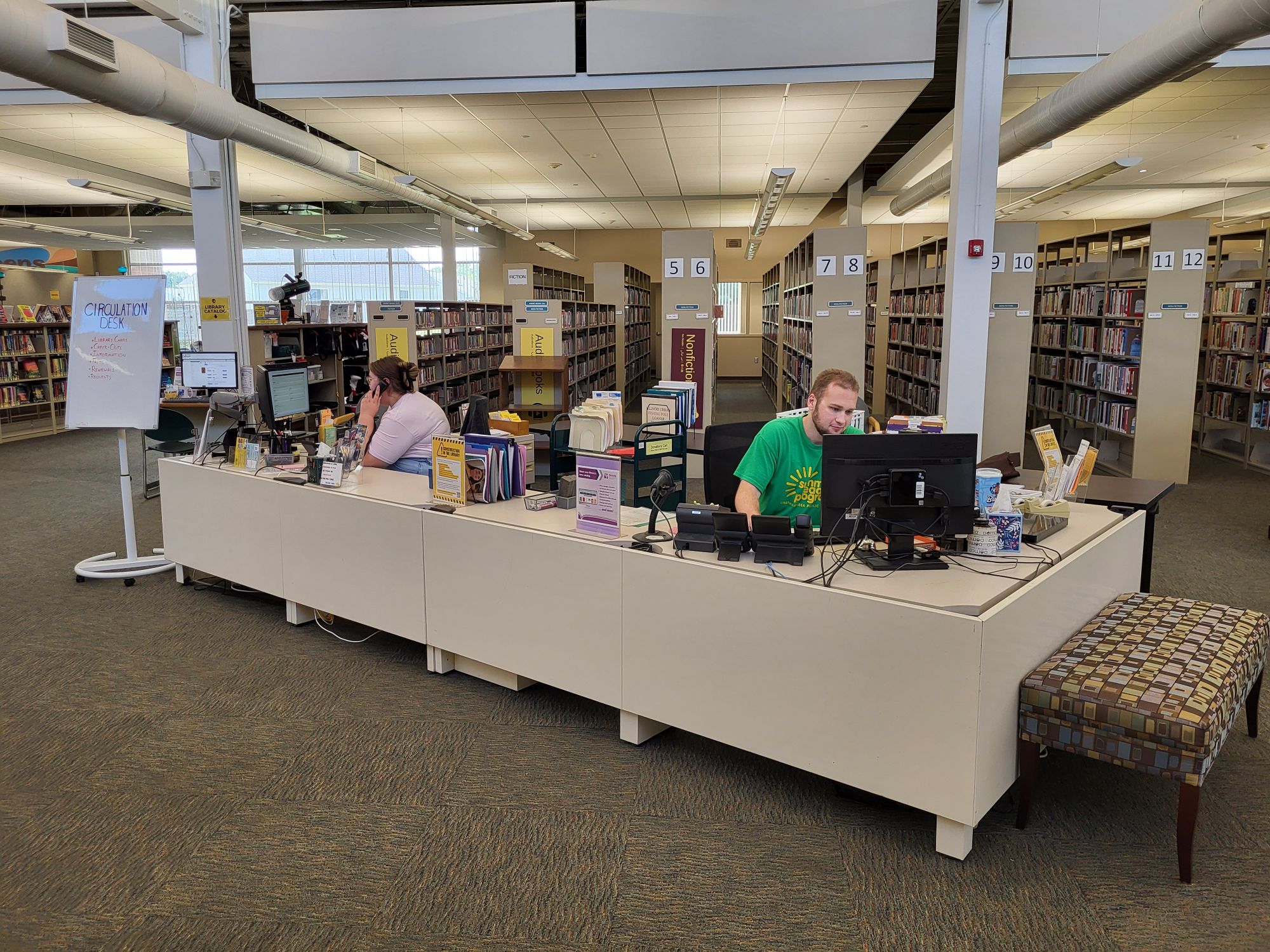 The new circulation desk: a long, rectangular desk with a computer on two front-facing corners. A staff member is seated at each station.