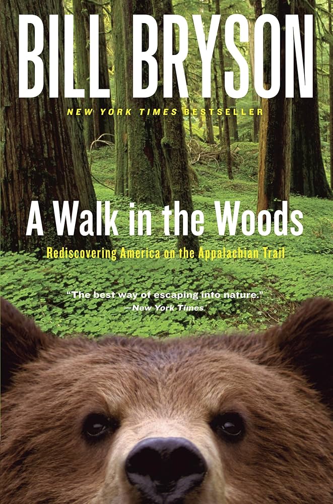 Book cover of A Walk in the Woods by Bill Bryson. On the bottom half of the cover, there is the face of a brown bear looking at you. In the background it shows a forest with tree trunks and a bunch of green weeds on the ground.